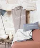 A collection of soft home textiles, including pillows and blankets, arranged on a shelf and a sofa, with a lamp and side table in the foreground. Produktbild-Vorschau