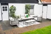 An outdoor patio set with a sofa, chairs, table, and bench, set against a white brick wall with a door and window. Produktbild-Vorschau