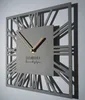 A modern square wall clock with Roman numerals and a geometric frame, casting a shadow on a light gray wall. Náhled obrázku produktu
