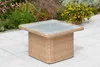 A square wicker coffee table with a glass top sits on a paved patio, with plants in the background. Produktbild-Vorschau