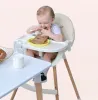 A baby is eating in a high chair next to a table with milk and bread. Produktbild-Vorschau