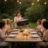 A group of people are enjoying an outdoor dinner party in the evening, with string lights overhead. One woman is presenting a black cooler box to the group. Produktbild-Vorschau