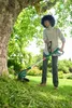 A person is using a cordless grass trimmer in a garden next to a large tree. Produktbild-Vorschau
