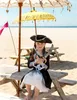 A young girl dressed as a pirate sits on a picnic bench on a sandy beach, holding two toy swords. Produktbild-Vorschau