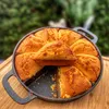 Cornbread baked in a cast iron pan, placed on a wooden cutting board, with a blurred green background of foliage. Produktbild-Vorschau