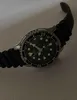 A close-up shot of a men's wristwatch with a black strap and a black dial. The watch face is illuminated, showing the hands and hour markers. Produktbild-Vorschau
