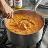 A hand is stirring a pot of creamy orange stew on a stovetop, with two jars of spices in the background. Produktbild-Vorschau