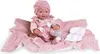 A baby doll dressed in pink, sitting on a pink blanket with a bottle and diapers. Anteprima dell’immagine prodotto