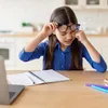 A young girl wearing glasses is sitting at a table, rubbing her eyes while looking at a laptop. There is a notebook and pens on the table. Voir les images du produit