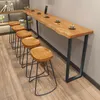 A long, narrow wooden bar table with a live edge, paired with several wooden stools, set against a light gray wall and wooden floor. Produktbild-Vorschau