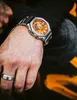 A close-up shot of a person's wrist wearing a silver watch with an orange face. Produktbild-Vorschau
