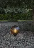 A decorative sphere light sits on a gravel path in a garden, surrounded by trees and bushes. Produktbild-Vorschau