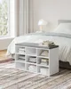 A white storage bench with a gray cushion and fabric items inside, placed in a bedroom next to a bed. Náhled obrázku produktu