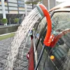 A portable camping shower is attached to the side of a red car, with water spraying from the showerhead. Náhled obrázku produktu