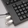 A close-up view of a black keyboard with two USB flash drives plugged into its side, and a USB cable connected to the keyboard. Náhled obrázku produktu