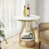 A round side table with a marble-like top and gold metal legs, placed next to a chair in a well-lit room with sheer curtains. Produktbild-Vorschau