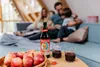 A family is relaxing on a couch in a cozy living room, with a bottle of juice and apples on a coffee table in the foreground. Produktbild-Vorschau
