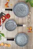 A set of three frying pans with speckled gray interiors and black handles are arranged on a wooden surface, surrounded by ingredients like cherry tomatoes, bell peppers, and leafy greens. Produktbild-Vorschau