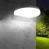 A modern, circular LED wall lamp is mounted on a textured concrete wall, illuminating the surrounding area and a lush green lawn with trees in the background at dusk. Podgląd zdjęcia produktu