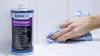 A hand is wiping a white tiled surface with a blue microfiber cloth, next to a can of Beko Allclean surface cleaner. Produktbild-Vorschau