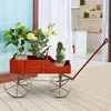 A red wooden wagon planter filled with various plants sits on a wooden deck, with a kitchen visible through a sliding glass door in the background. Podgląd zdjęcia produktu