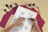 A person's hands are shown drawing on a paper with a cat design, with bottles of fabric paint and a paintbrush laid out on a pink shirt. Produktbild-Vorschau