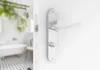 A close-up of a silver door handle and lock mechanism on a white door, with a blurred bathroom in the background. Produktbild-Vorschau