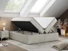 A beige boxspring bed with the storage compartment open, showing the mattress lifted by hydraulic arms. The bed is in a room with skylights and a bedside table. Produktbild-Vorschau
