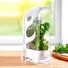 A clear plastic herb keeper with fresh herbs inside, placed on a wooden surface next to a cutting board with more herbs and a knife. Produktbild-Vorschau