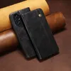 A black leather phone case is shown in a close-up shot, resting on a textured brown surface. The case is open, revealing the camera array of a phone. Rolls of leather are visible in the background. Produktbild-Vorschau