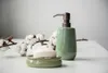 A green ceramic soap dispenser and a bar of soap on a soap dish, with a branch of cotton on a white surface. Produktbild-Vorschau