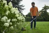 A man is using a cordless grass trimmer in a garden next to a large bush with white flowers. Produktbild-Vorschau