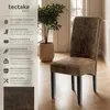 A brown upholstered dining chair with black legs is shown in a room with a plant and a window. Náhled obrázku produktu