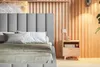 A close-up view of a bed with a grey upholstered headboard and beige bedding, set against a wood-paneled wall. Náhled obrázku produktu