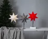 Three star-shaped lamps are displayed on a table in front of a Christmas tree and a gray wall. Podgląd zdjęcia produktu