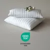 Two white pillows are stacked on a light gray surface, with a small bouquet of white flowers on top of the upper pillow. Below the pillows, there is a green and white logo with text. Produktbild-Vorschau