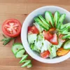 A salad with tomatoes and cucumbers is presented on a wooden surface, with a sliced tomato and cucumber spiral on the side. Anteprima dell’immagine prodotto