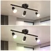 A black LED ceiling light fixture with three adjustable spotlights is shown mounted on a white ceiling. The image is split horizontally, with the top half showing the lights emitting a warm white light and the bottom half showing them emitting colored ligh Produktbild-Vorschau