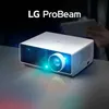 A white LG ProBeam projector is shown on a reflective surface, emitting a blue light. A person is visible in the background holding a tablet. Náhled obrázku produktu