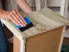 A person is using a blue and black squeegee to apply a wood-grain adhesive film to the surface of a piece of furniture. Produktbild-Vorschau