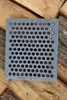 A square cast iron grate with many circular holes is placed on a pile of wood. Voir les images du produit