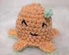 A crocheted toy with a face and small arms and legs, sitting on a white fluffy surface. Náhľad obrázku produktu