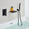 A black built-in bathtub faucet with a waterfall spout and a handheld showerhead is shown mounted on a white wall above a bathtub filled with water. Podgląd zdjęcia produktu