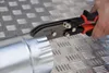 A hand is using a pair of metal shears to cut a metal pipe on a textured metal surface. Anteprima dell’immagine prodotto
