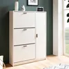 A white shoe cabinet with three flip drawers and a closed cabinet door, placed in a room with a dark green wall and a window. Produktbild-Vorschau