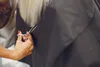A close-up of a person's hand using scissors to cut blonde hair, with a black cape draped around the hair. Anteprima dell’immagine prodotto