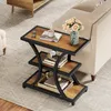 A three-tiered side table with a rustic brown wood finish and black metal frame, placed next to a sofa in a living room setting. It is decorated with a vase of flowers, books, and a pair of glasses. Produktbild-Vorschau