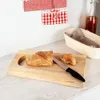 A wooden cutting board with bread and a knife on a countertop, with decorative boxes and a bottle in the background. Náhľad obrázku produktu