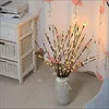 Decorative LED branches in a vase, placed next to a white cabinet with woven baskets, with a flamingo-patterned curtain in the background. Náhled obrázku produktu