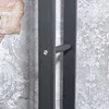 A close-up view of a metal handle on a dark gray frame, set against a textured background with a brick pattern on the left and a floral damask pattern on the right. Podgląd zdjęcia produktu